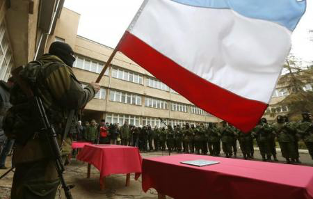 Um homem armado, tido como um soldado russo, segura uma bandeira da Crimeia enquanto membros de uma unidade pró-Rússia de autodefesa ficam em formação antes de dar um juramento ao governo da Crimeia, em Simferopol.